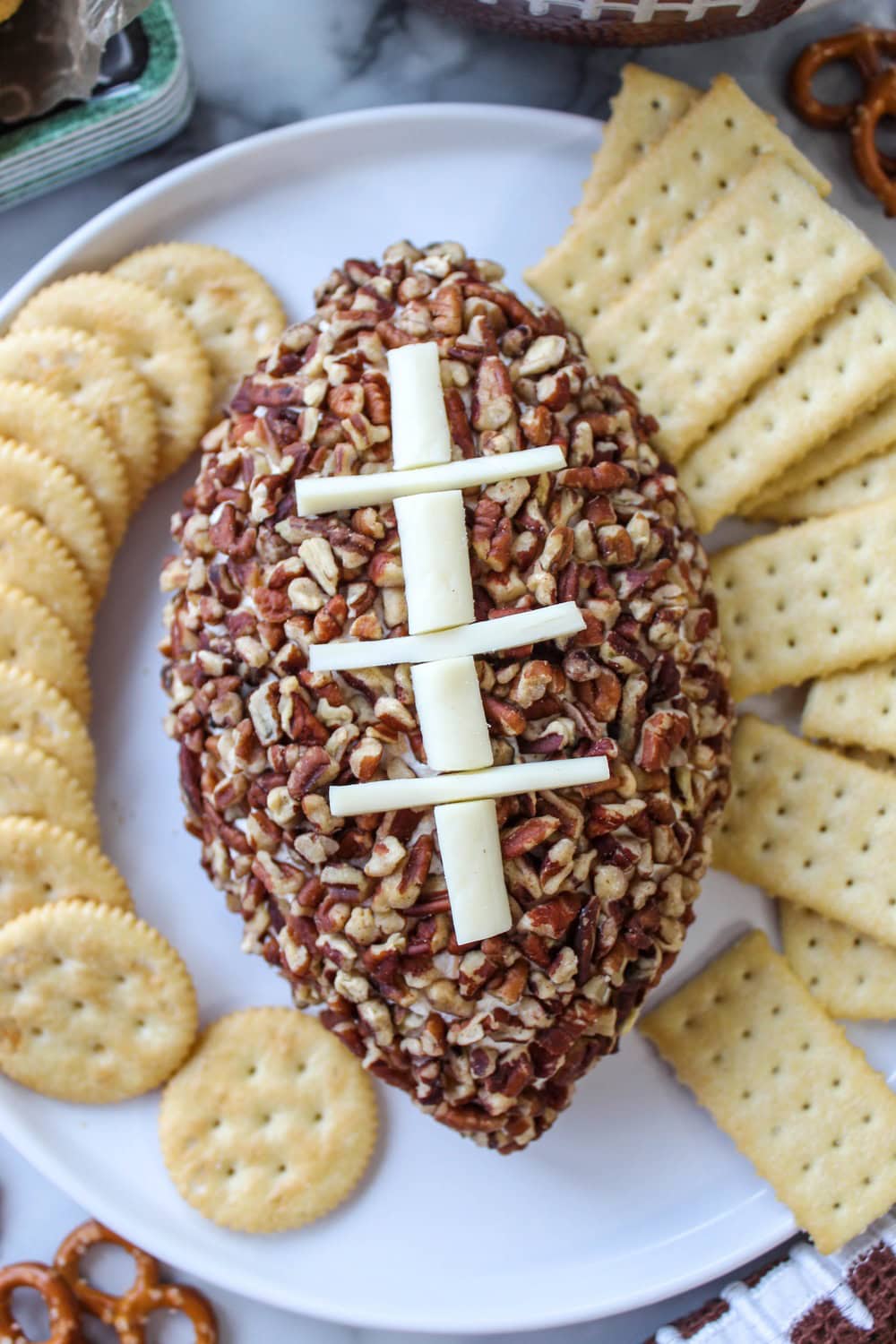 A football-shaped cheeseball coated with nuts and decorated with white cheese laces, served on a platter with crackers